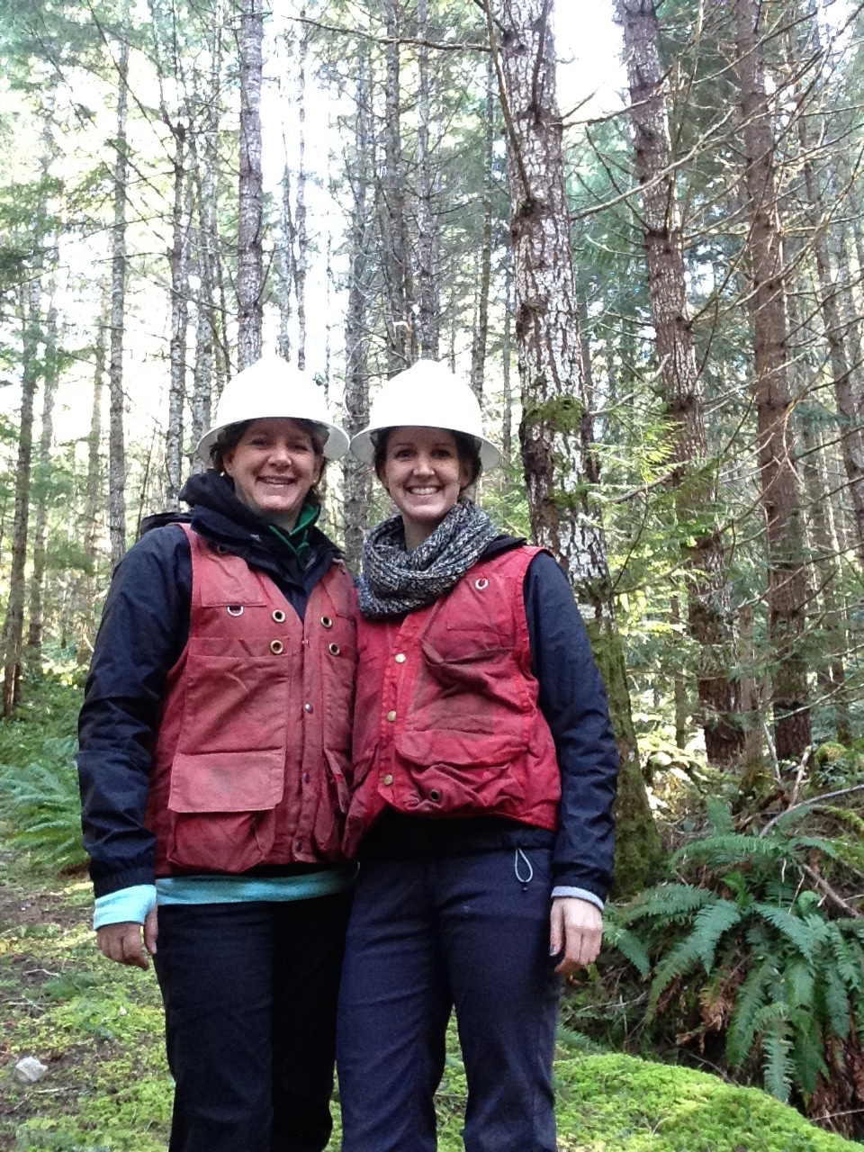 Two women smiling the the camera while wearing white hard hats and bright red vests. They are standing in a forest.